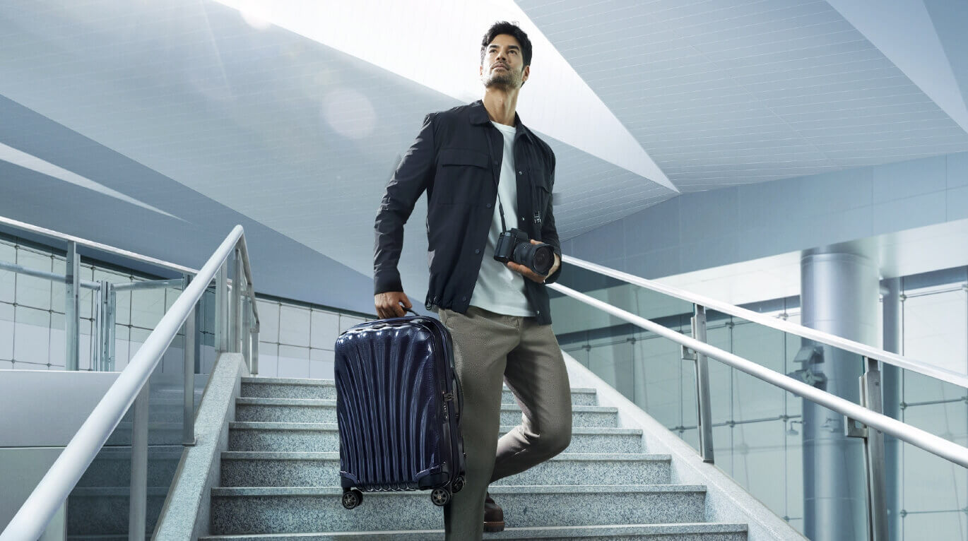 Male carrying the C-LITE luggage with his right hand, slinged camera held in left hand, posing on the stairs looking up ahead